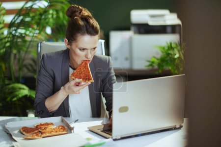 Foto de Lugar de trabajo sostenible. moderna mujer de mediana edad trabajadora en un traje de negocios gris en la moderna oficina verde con pizza y portátil. - Imagen libre de derechos