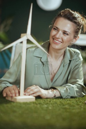 Foto de Eco Negocios. sonriente elegante propietaria de una pequeña empresa mujer en blusa verde en moderna oficina verde con molino de viento - Imagen libre de derechos