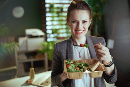 Téléchargez les photos : Un lieu de travail durable. souriant femme propriétaire de petite entreprise moderne dans un costume d'affaires gris dans un bureau vert moderne avec ordinateur portable manger salade. - en image libre de droit