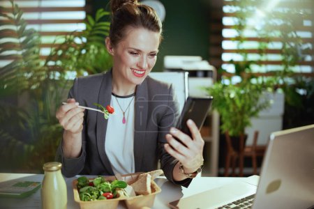 Photo for Sustainable workplace. happy modern middle aged small business owner woman in a grey business suit in modern green office with laptop and smartphone eating salad. - Royalty Free Image