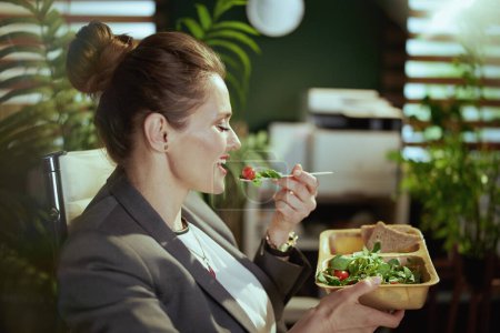 Téléchargez les photos : Un lieu de travail durable. souriant femme d'âge moyen moderne travailleur dans un costume d'affaires gris dans le bureau vert moderne manger de la salade. - en image libre de droit