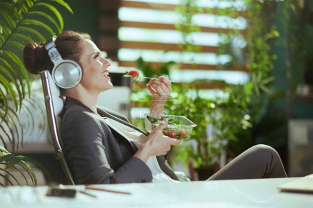 Téléchargez les photos : Un lieu de travail durable. souriant femme comptable d'âge moyen moderne dans un costume d'affaires gris dans le bureau vert moderne manger de la salade et écouter la musique avec écouteurs. - en image libre de droit
