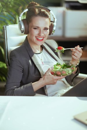 Téléchargez les photos : Un lieu de travail durable. souriant femme d'âge moyen moderne travailleur dans un costume d'affaires gris dans le bureau vert moderne avec écouteurs manger de la salade. - en image libre de droit