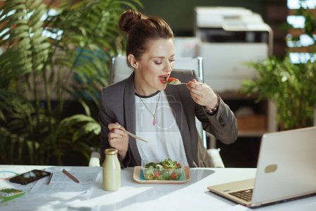 Foto de Lugar de trabajo sostenible. moderna mujer propietaria de una pequeña empresa en un traje de negocios gris en la oficina verde moderna con portátil comer ensalada. - Imagen libre de derechos