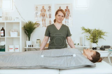 Photo for Healthcare time. smiling female massage therapist in massage cabinet with client doing whiskey swaddling procedure in thermal blanket. - Royalty Free Image