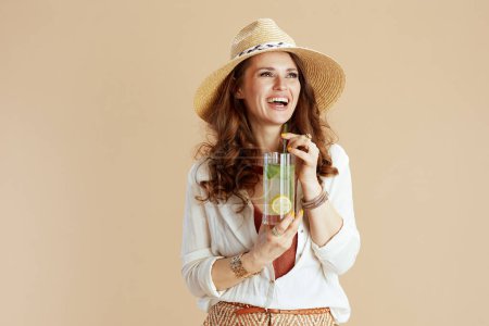 Téléchargez les photos : Vacances à la plage. femme élégante et souriante en chemisier blanc et short isolé sur fond beige avec cocktail et chapeau d'été. - en image libre de droit