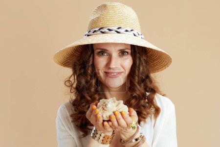 Téléchargez les photos : Vacances à la plage. femme élégante et souriante de 40 ans en chemisier blanc et short isolé sur fond beige avec coquillage et chapeau de paille. - en image libre de droit