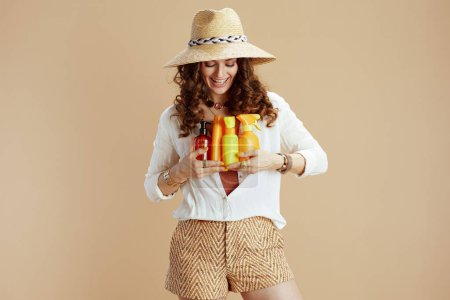 Photo for Beach vacation. smiling elegant middle aged housewife in white blouse and shorts isolated on beige with spf and summer hat. - Royalty Free Image