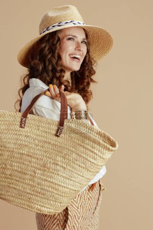 Téléchargez les photos : Vacances à la plage. heureuse femme au foyer moderne en chemisier blanc et short isolé sur beige avec sac en paille et chapeau en paille. - en image libre de droit