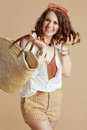 Photo for Beach vacation. smiling modern housewife in white blouse and shorts isolated on beige with straw bag and sunglasses. - Royalty Free Image