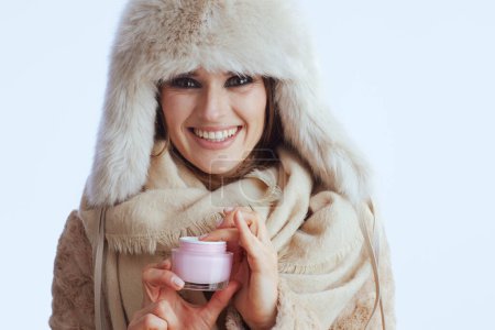 Téléchargez les photos : Femme élégante souriante en manteau d'hiver et chapeau de fourrure isolé sur blanc avec pot cosmétique. - en image libre de droit