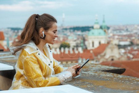 Photo for Relaxed young woman in yellow blouse and raincoat in Prague Czech Republic with headphones using smartphone against city panorama. - Royalty Free Image