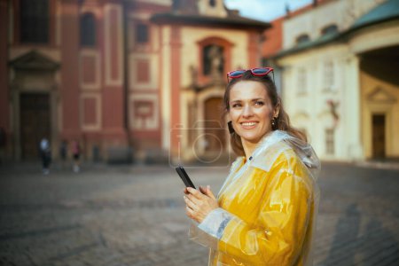 Foto de Mujer joven feliz en blusa amarilla y impermeable en Praga República Checa utilizando el teléfono inteligente. - Imagen libre de derechos