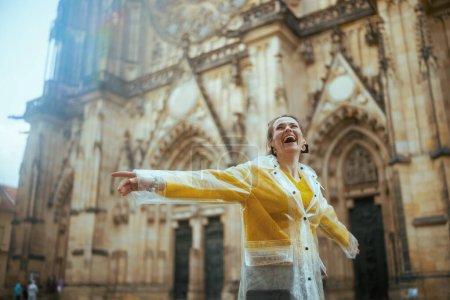 Téléchargez les photos : Heureuse voyageuse d'âge moyen à la mode en chemisier jaune et imperméable à Prague République tchèque profiter de la promenade. - en image libre de droit