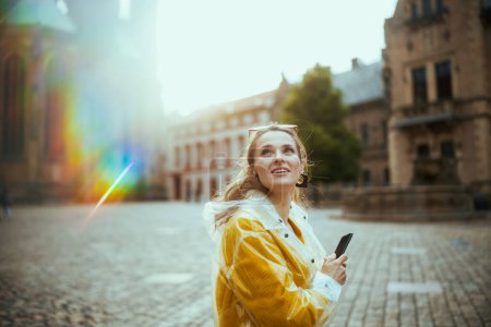Téléchargez les photos : Heureuse jeune voyageuse en chemisier jaune et imperméable à Prague République tchèque avec smartphone visites. - en image libre de droit
