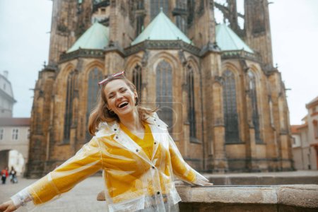 Foto de Sonriente mujer moderna en blusa amarilla y impermeable en Praga República Checa teniendo excursión. - Imagen libre de derechos