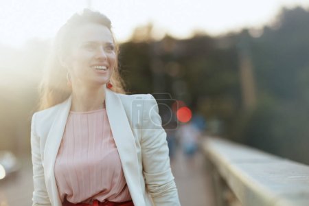 Photo for Happy trendy middle aged woman in pink dress and white jacket in the city on the bridge. - Royalty Free Image