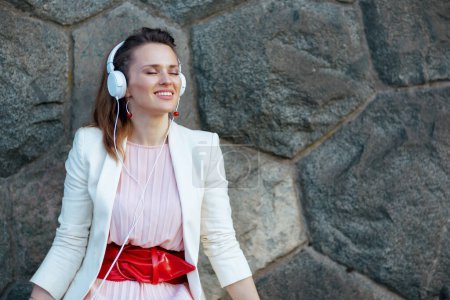 Téléchargez les photos : Jeune femme souriante en robe rose et veste blanche dans la ville à l'écoute de la musique avec écouteurs contre mur de pierre. - en image libre de droit