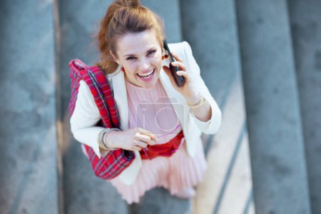 Foto de Vista superior de la feliz mujer moderna de 40 años en vestido rosa y chaqueta blanca en la ciudad con bolsa roja hablando en un teléfono inteligente. - Imagen libre de derechos