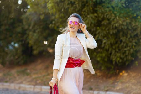 Foto de Mujer elegante sonriente en vestido rosa y chaqueta blanca en la ciudad con gafas de sol caminando. - Imagen libre de derechos
