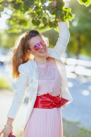 Téléchargez les photos : Femme élégante et souriante en robe rose et veste blanche dans la ville avec des lunettes de soleil. - en image libre de droit