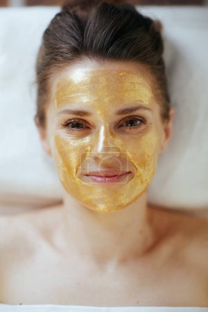 Foto de Tiempo de atención médica. Vista superior de la mujer moderna relajada en el gabinete de masaje con máscara cosmética dorada en la cara que pone en la mesa de masaje. - Imagen libre de derechos