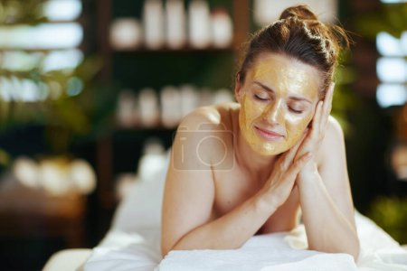 Photo for Healthcare time. relaxed modern middle aged woman in spa salon with golden cosmetic mask on face laying on massage table. - Royalty Free Image