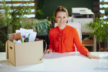 Photo for New job. smiling modern woman worker in modern green office in red blouse with personal belongings in cardboard box. - Royalty Free Image