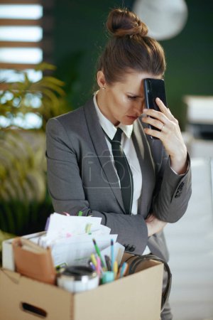 Photo for New job. unhappy modern female employee in modern green office in grey business suit with personal belongings in cardboard box and smartphone. - Royalty Free Image
