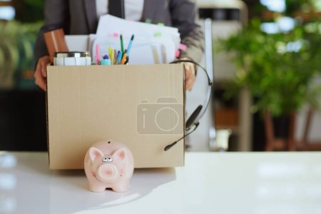 Photo for New job. Closeup on modern 40 years old woman employee in modern green office with personal belongings in cardboard box and piggy bank. - Royalty Free Image