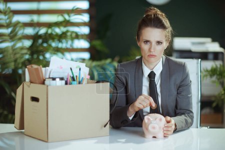 Photo for New job. concerned modern 40 years old woman employee in modern green office in grey business suit with personal belongings in cardboard box putting coin into piggy bank. - Royalty Free Image