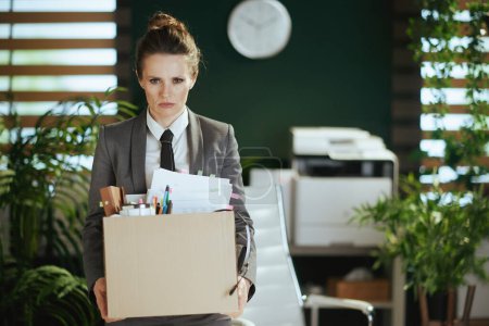 Foto de Un nuevo trabajo. trabajadora moderna preocupada en moderna oficina verde en traje de negocios gris con pertenencias personales en caja de cartón. - Imagen libre de derechos