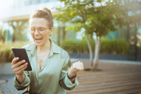 Téléchargez les photos : Heureuse femme d'affaires moderne d'âge moyen dans le quartier des affaires en chemisier vert et lunettes avec smartphone se réjouissant. - en image libre de droit