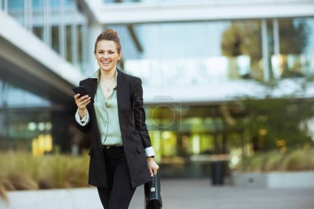 Foto de Sonriente moderna empleada de 40 años cerca del centro de negocios en chaqueta negra con teléfono inteligente y maletín caminando. - Imagen libre de derechos