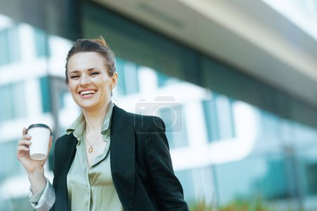Foto de Retrato de mujer trabajadora moderna de mediana edad sonriente en el distrito de negocios con chaqueta negra con taza de café caminando. - Imagen libre de derechos