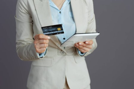 Téléchargez les photos : Gros plan sur femme d'affaires d'âge moyen dans un costume d'affaires léger avec carte de crédit en utilisant tablette PC sur fond gris. - en image libre de droit