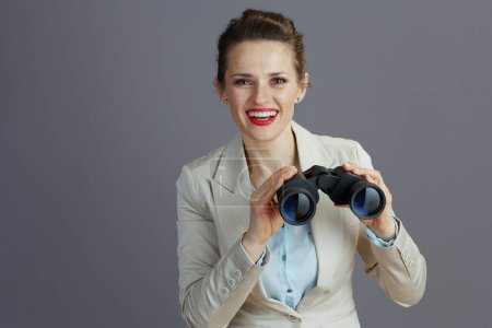 Photo for Happy modern middle aged small business owner woman in a light business suit with binoculars isolated on grey background. - Royalty Free Image