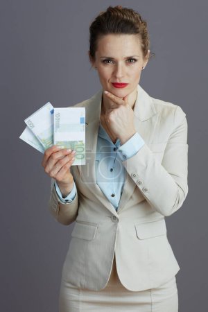 Photo for Pensive trendy 40 years old woman worker in a light business suit with euros money packs isolated on gray. - Royalty Free Image