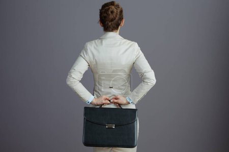 Foto de Visto desde atrás mujer propietaria de una pequeña empresa en un traje de negocios ligero con maletín contra fondo gris. - Imagen libre de derechos