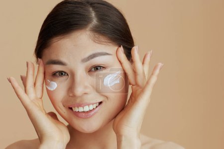 Foto de Retrato de mujer joven asiática con crema facial en la cara sobre fondo beige. - Imagen libre de derechos