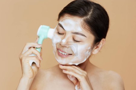 Foto de Retrato de joven mujer asiática con masajeador lavando la cara sobre fondo beige. - Imagen libre de derechos