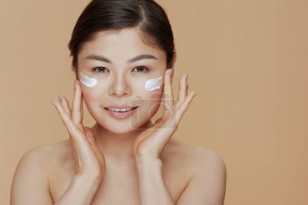 Téléchargez les photos : Portrait de femme moderne avec crème faciale sur le visage sur fond beige. - en image libre de droit