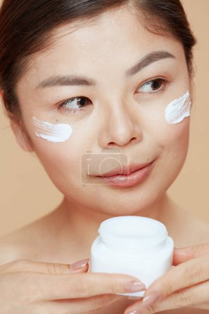 Foto de Mujer joven con frasco de crema facial y crema facial en la cara aislada sobre fondo beige. - Imagen libre de derechos
