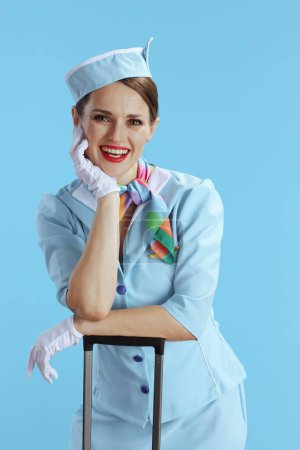 Téléchargez les photos : Heureuse élégante hôtesse de l'air femme sur fond bleu en uniforme bleu avec sac de voyage. - en image libre de droit
