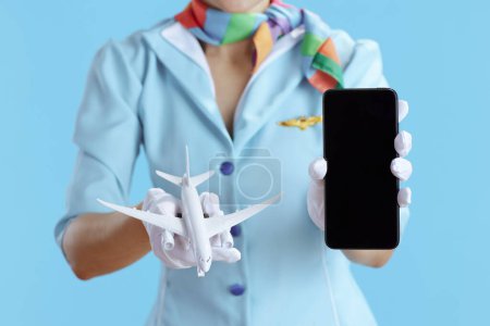 Photo for Closeup on flight attendant woman on blue background in blue uniform with a little airplane showing smartphone blank screen. - Royalty Free Image