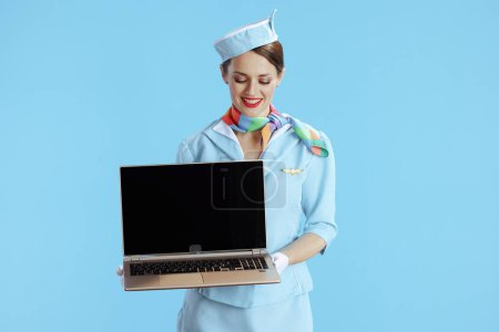 Téléchargez les photos : Heureuse élégante hôtesse de l'air isolé sur fond bleu en uniforme bleu montrant écran blanc ordinateur portable. - en image libre de droit