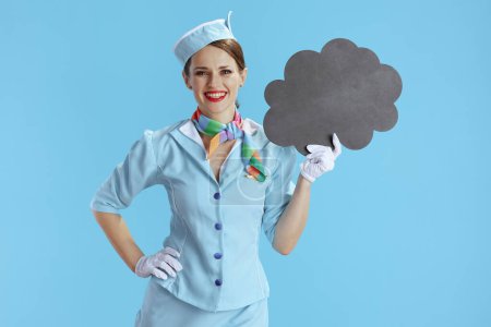 Foto de Asistente de vuelo femenino elegante feliz contra fondo azul en uniforme azul que muestra el tablero en blanco de la forma de la nube. - Imagen libre de derechos