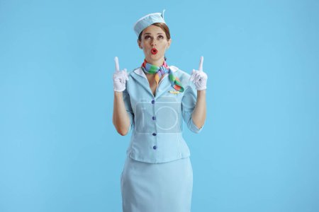 Foto de Sorprendida azafata femenina elegante sobre fondo azul en uniforme azul apuntando hacia arriba en el espacio de copia. - Imagen libre de derechos