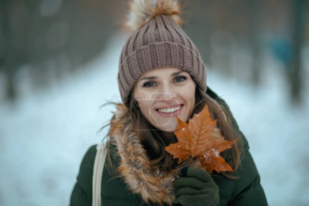 Téléchargez les photos : Portrait de femme moderne souriante en manteau vert et chapeau brun à l'extérieur dans le parc de la ville en hiver avec mitaines, feuille d'automne et chapeau de bonnet. - en image libre de droit