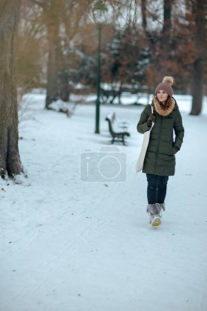 Téléchargez les photos : Portrait complet de femme moderne coûteuse en manteau vert et chapeau brun à l'extérieur dans le parc de la ville en hiver avec bonnet chapeau. - en image libre de droit